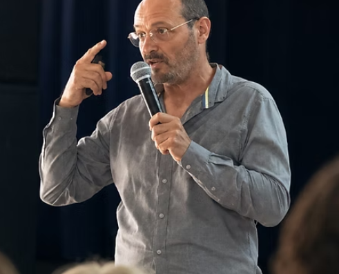 Un homme d'âge mûr en chemise grise s'exprime au micro lors d'une conférence professionnelle.